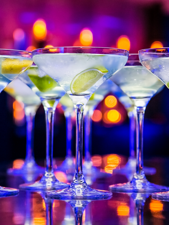 A row of martini glasses sitting on top of a bar. The martini glasses are lined up in a row, and they are all different shapes and sizes. The martini glasses are all different colors and sizes, and they are all sitting on top of a bar.