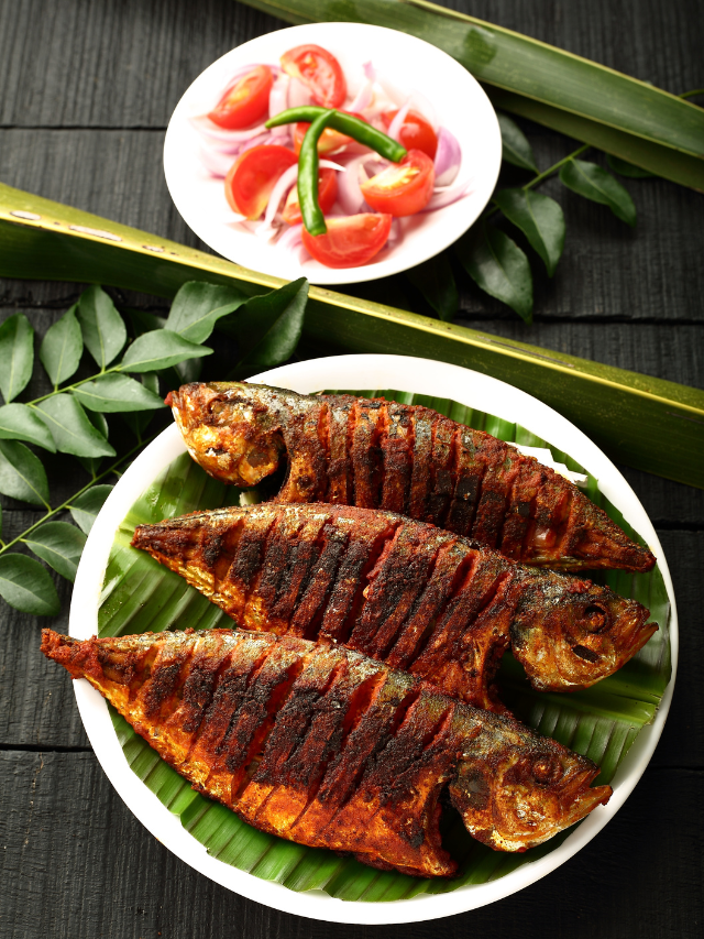 A plate of fried fish on a banana leaf with a side of tomatoes and onions. The fish is a type of Bangda, which is a popular fish in India. The image is relevant to the topic of seafood dishes you should try at sea beaches.