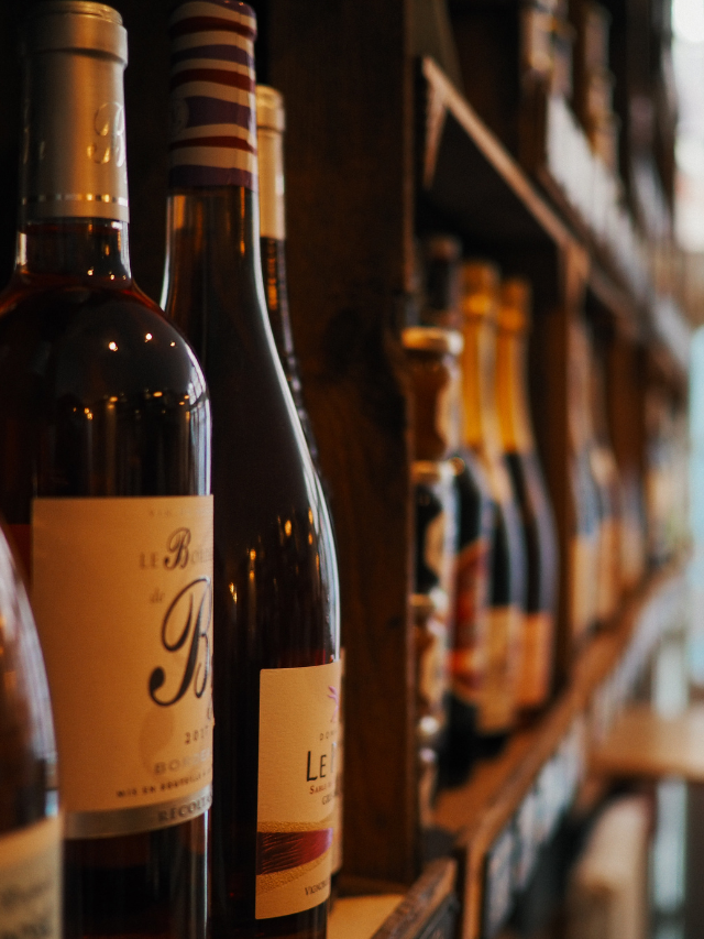 A row of wine bottles sitting on top of each other on a wooden shelf in a store. The bottles are all different colors and shapes, and the shelf is made of wood. The bottles are all different shapes and sizes, and the shelf is made of wood.